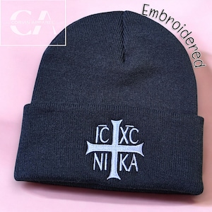 May include: A dark gray knitted beanie with a white embroidered cross and the text "IC XC NI KA". The beanie is embroidered with the text "Embroidered" in the top right corner.