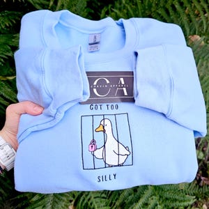 Puede incluir: Sudadera azul claro con un dibujo de un pato blanco sosteniendo un bolso rosa dentro de una jaula. El texto "GOT TOO SILLY" está debajo. La sudadera tiene el logo "CORVIN APPAREL" en la etiqueta.