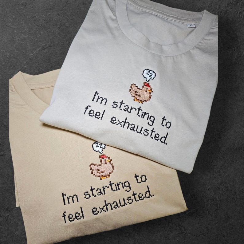Op de afbeelding: Twee gevouwen t-shirts, &eacute;&eacute;n beige en &eacute;&eacute;n lichtgrijs, zijn op elkaar gestapeld. Elk shirt toont de tekst "I'm starting to feel exhausted" met een gepixelde kippengrafiek en een slaapbel.