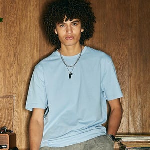 Op de afbeelding: Een lichtblauw T-shirt met korte mouwen. Het shirt wordt gecombineerd met een grijze broek en een zilveren ketting met een zwarte hanger. De achtergrond is een houten muur.