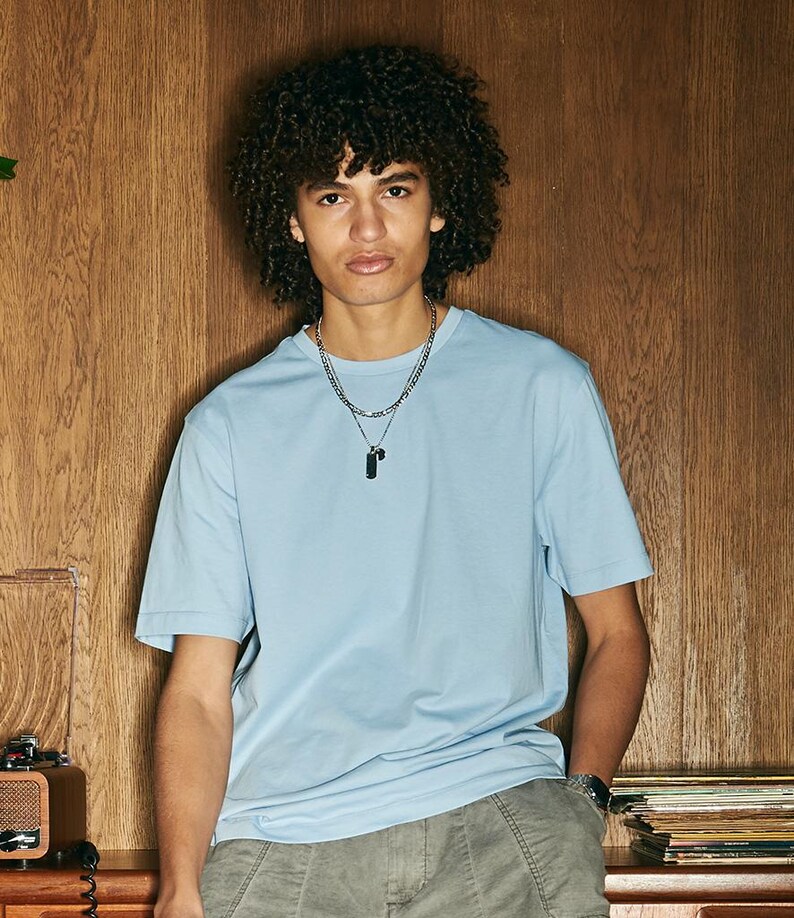 Op de afbeelding: Een lichtblauw T-shirt met korte mouwen. Het shirt wordt gecombineerd met een grijze broek en een zilveren ketting met een zwarte hanger. De achtergrond is een houten muur.