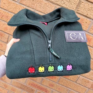 May include: Dark green fleece pullover with a zip-up collar. The pullover features a colorful pixelated design of five small characters. A tag that says "Corvin Apparel" is on the upper right side.