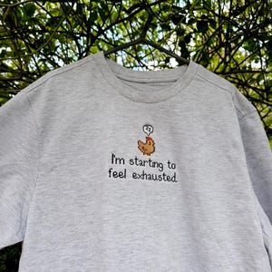 Op de afbeelding: Een lichtgrijs sweatshirt met een ronde hals. Het shirt heeft een geborduurd ontwerp van een bruine kip met een denkbel en de tekst "I'm starting to feel exhausted."