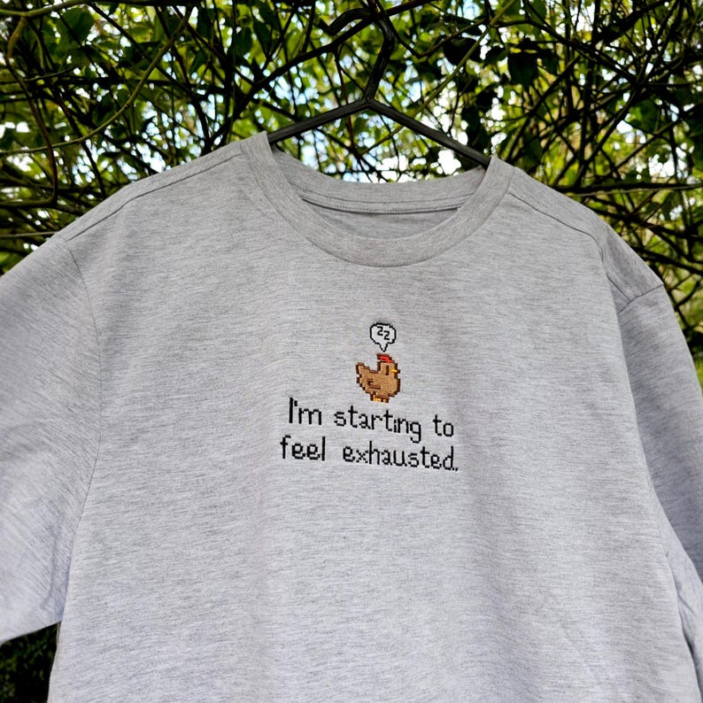 Op de afbeelding: Een lichtgrijs sweatshirt met een ronde hals. Het shirt heeft een geborduurd ontwerp van een bruine kip met een denkbel en de tekst "I'm starting to feel exhausted."