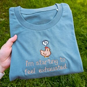 Op de afbeelding: Een tealkleurig T-shirt met een gepixeld kippenontwerp en de tekst "I'm starting to feel exhausted." Het ontwerp bevat een tekstballon met "zz" erin. Het shirt is opgevouwen en wordt door een hand vastgehouden.