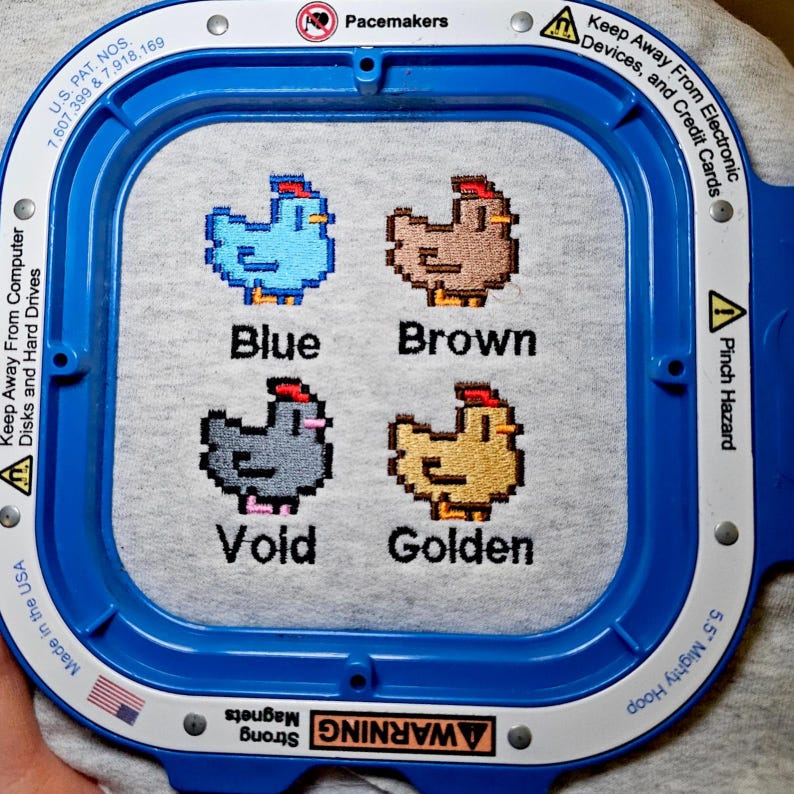 Op de afbeelding: Een grijze stof met vier gepixelde kippenontwerpen in blauw, bruin, void en goud. De naam van elke kleur staat onder elke kip. De stof wordt in een blauwe borduurring gehouden.