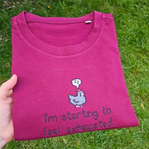 Op de afbeelding: Een bordeauxrode t-shirt met ronde hals. De gevouwen t-shirt heeft een pixelafbeelding van een slapende vogel en de tekst "I'm starting to feel exhausted." De t-shirt wordt door een hand vastgehouden.