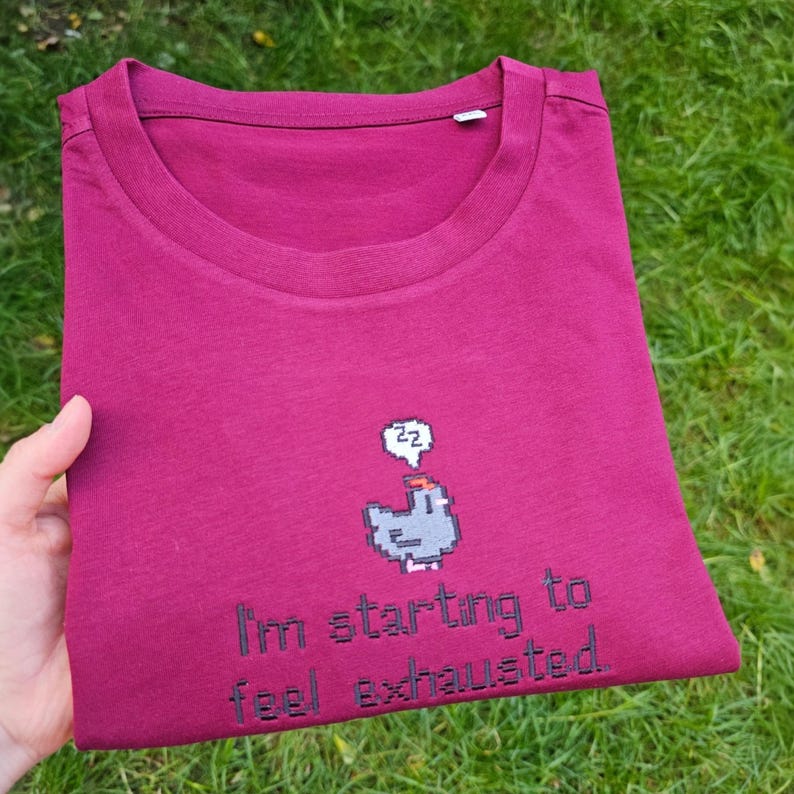 Op de afbeelding: Een bordeauxrode t-shirt met ronde hals. De gevouwen t-shirt heeft een pixelafbeelding van een slapende vogel en de tekst "I'm starting to feel exhausted." De t-shirt wordt door een hand vastgehouden.
