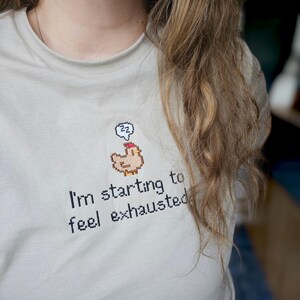Op de afbeelding: Een lichtgrijs t-shirt met de tekst "I'm starting to feel exhausted" in het zwart. Boven de tekst staat een gepixeld beeld van een kip met een gedachtewolk met "zz". Het shirt is gemaakt van zacht materiaal.