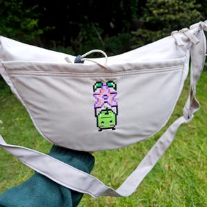 May include: Beige crossbody bag featuring a pixelated design of a purple flower and a green creature. The bag has a curved shape, a long adjustable strap, and a front pocket. The bag is made of a soft, durable material.