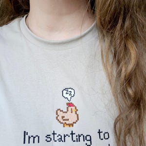 Op de afbeelding: Lichtbeige t-shirt met de tekst "I'm starting to feel exhausted." en een gepixelde kippengrafiek met een denkbel. De denkbel bevat de letters "zz".