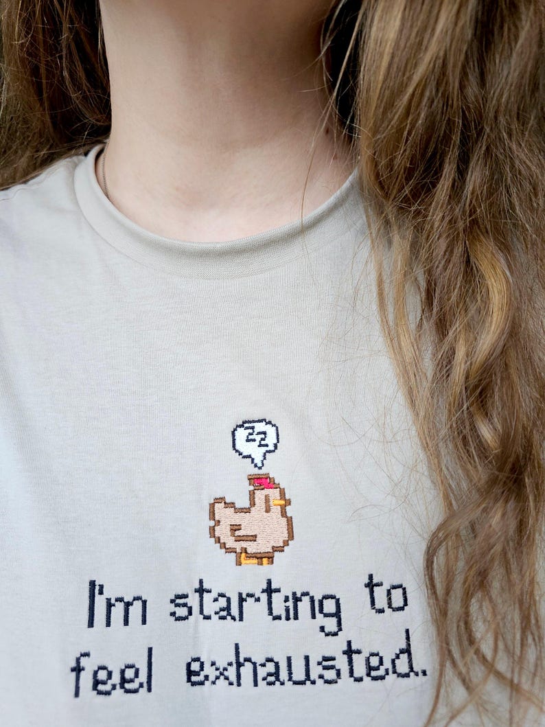 Op de afbeelding: Lichtbeige t-shirt met de tekst "I'm starting to feel exhausted." en een gepixelde kippengrafiek met een denkbel. De denkbel bevat de letters "zz".