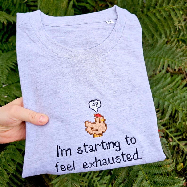 Op de afbeelding: Lichtgrijs T-shirt met een ronde hals, met een gepixeld kippenontwerp en de tekst "I'm starting to feel exhausted." Het shirt is opgevouwen en wordt tegen een achtergrond van groen gebladerte gehouden.