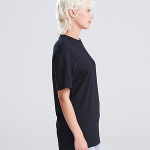 Op de afbeelding: Een zijkant van een zwart T-shirt met ronde hals. De korte mouwen worden gedragen door een persoon met kort, licht haar. Het shirt lijkt gemaakt van een zacht materiaal en heeft een eenvoudig, klassiek ontwerp.