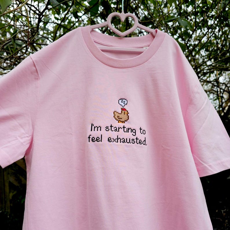 Op de afbeelding: Lichtroze T-shirt met een ronde hals en korte mouwen. Het shirt heeft een geborduurd ontwerp van een gepixeleerde kip met een denkbel en de tekst "I'm starting to feel exhausted."
