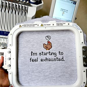 Op de afbeelding: Een grijs t-shirt in een borduurring met de tekst "I'm starting to feel exhausted" en een gepixeleerde kip. Een Brother borduurmachine en scherm zijn op de achtergrond.