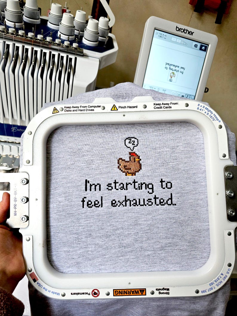 Op de afbeelding: Een grijs t-shirt in een borduurring met de tekst "I'm starting to feel exhausted" en een gepixeleerde kip. Een Brother borduurmachine en scherm zijn op de achtergrond.