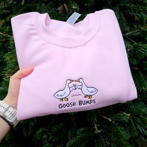 Puede incluir: Una sudadera rosa claro con cuello redondo. La sudadera presenta un diseño bordado de dos gansos blancos enfrentados, con el texto "Goose Bumps" debajo. El fondo es una vista borrosa de follaje verde.