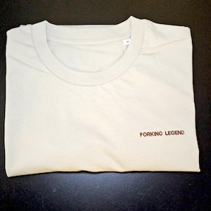 May include: A light beige crew neck t-shirt with short sleeves and a size M label at the inner collar. The front of the shirt features the text FORKING LEGEND embroidered in small brown capital letters on the lower right side of the chest area.