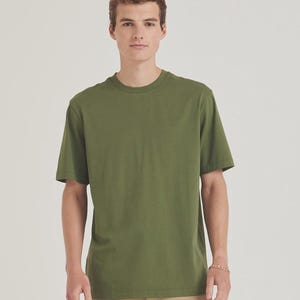 Op de afbeelding: Olijfgroen T-shirt met ronde hals. Het shirt met korte mouwen is effen van kleur en heeft een klassiek, casual design. Het model draagt een kaki broek en een gouden armband.