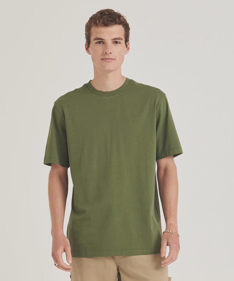 Op de afbeelding: Olijfgroen T-shirt met ronde hals. Het shirt met korte mouwen is effen van kleur en heeft een klassiek, casual design. Het model draagt een kaki broek en een gouden armband.