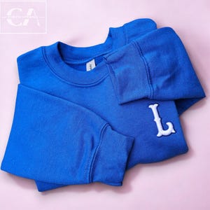 以下が含まれることがあります： 胸の右側に白い刺繍の文字Lが施されたロイヤルブルーのスウェットシャツです。