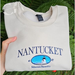 May include: A beige sweatshirt featuring the embroidered word "NANTUCKET" in navy blue. Below is a blue oval with a lighthouse illustration. The text "Massachusetts" is below. The sweatshirt is folded.