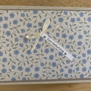 Könnte beinhalten: Rechteckige Grußkarte mit floralem Muster aus blauen Blumen und grünen Ranken auf cremefarbenem Hintergrund. Die Karte hat einen beigen Rand und ein cremefarbenes Band mit dem Text "Zum Geburtstag".