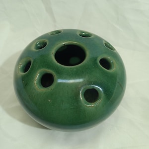 May include: A round, dark green ceramic flower frog with multiple holes for arranging stems. The frog has a glossy finish and a spherical shape, designed to hold flowers in place. The holes vary in size.