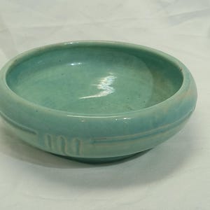 May include: A round, turquoise ceramic bowl with a slightly textured surface. The bowl has a wide rim and a series of three vertical indentations on the side. The bowl is a light blue-green color.