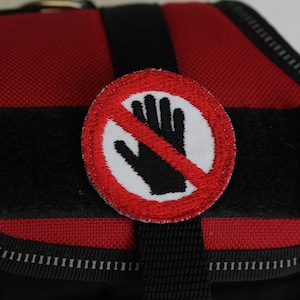 Könnte beinhalten: Rotes und schwarzes Hundegeschirr mit einem weißen Kreis und einer schwarzen Hand, die von einer roten Linie durchgestrichen ist. Der Kreis trägt den Text "No Hands".