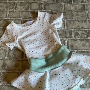May include: A white and mint green two-piece outfit for a child. The top is a short-sleeved shirt with a white background and a colorful sprinkle pattern. The bottom is a mint green skirt with a white ruffle and a colorful sprinkle pattern.