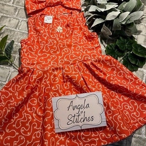 May include: A red sleeveless dress with a white pattern of connected loops. The dress has a gathered skirt and a matching red bow headband. The dress has a white tag with the text "Angela n' Stitches".