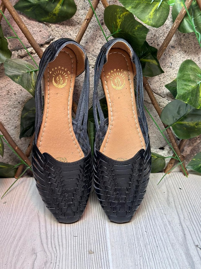 May include: A pair of black leather sandals with a woven design. The sandals have a pointed toe and a flat sole. The brand name "APACHE" is visible on the inside of each sandal.