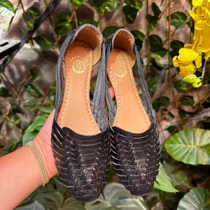 May include: A pair of black leather sandals with a woven design. The sandals have a pointed toe and a low heel. The sandals are made by the brand "Arapahoe".