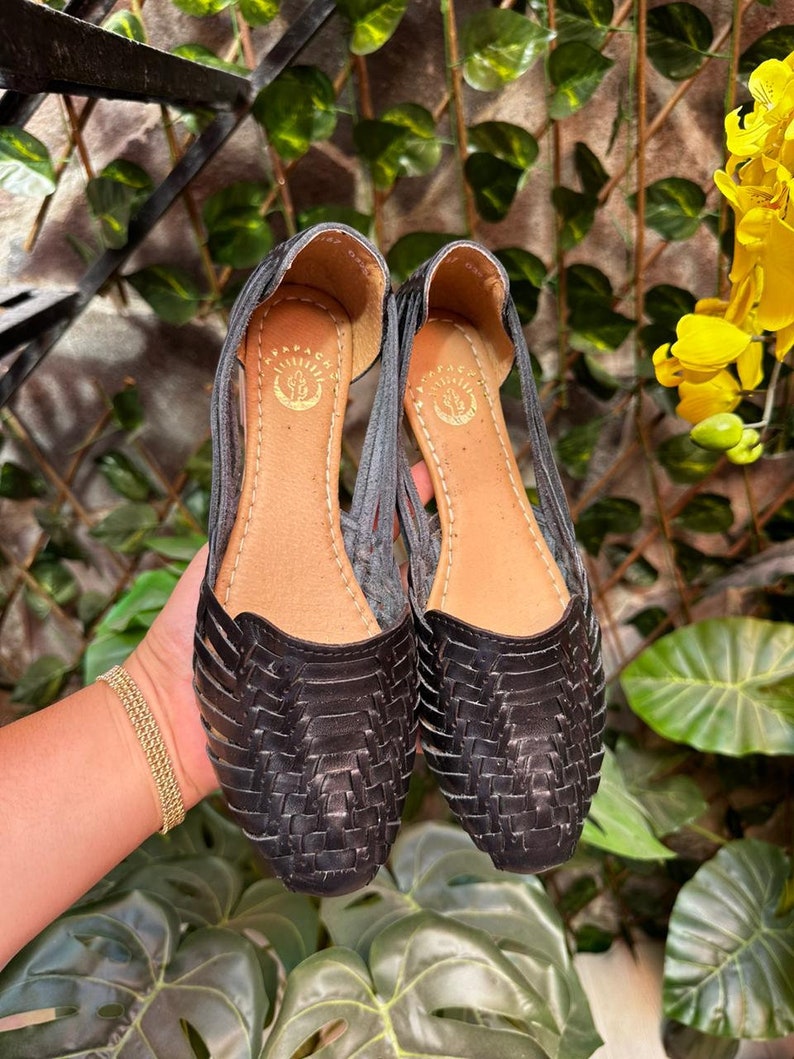 May include: A pair of black leather sandals with a woven design. The sandals have a pointed toe and a low heel. The sandals are made by the brand "Arapahoe".