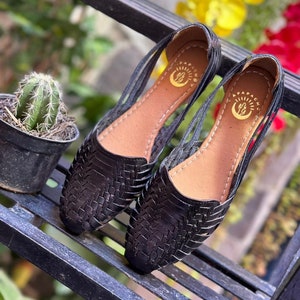 May include: A pair of black leather sandals with woven straps and a cutout design. The sandals have a brown leather insole and a gold logo that says "Apapacho".