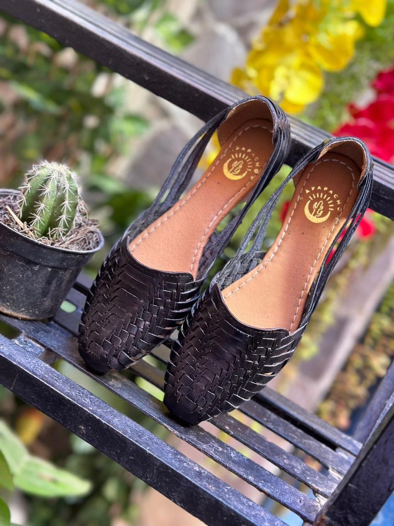 May include: A pair of black leather sandals with woven straps and a cutout design. The sandals have a brown leather insole and a gold logo that says "Apapacho".
