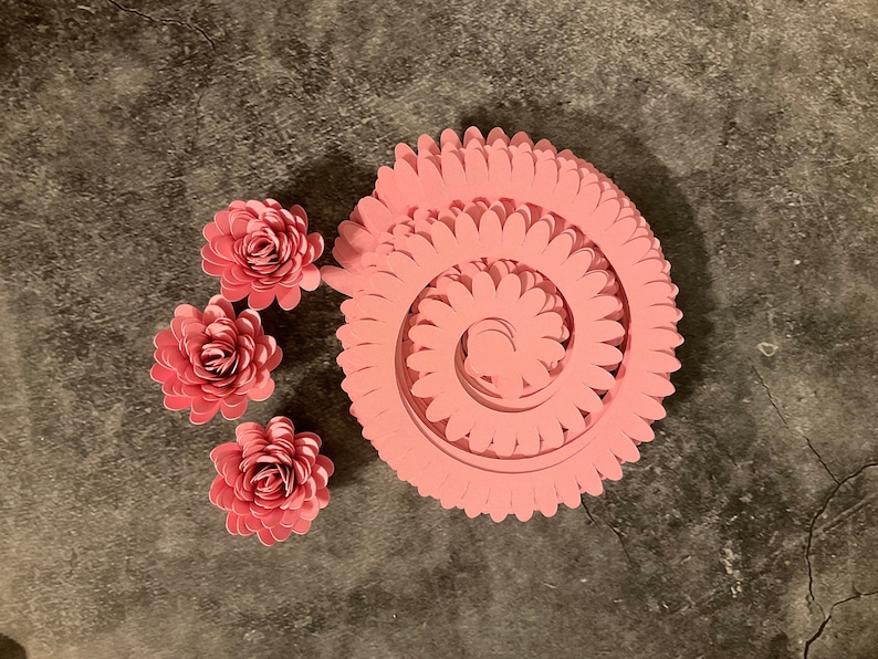 Pode incluir: Uma flor de papel rosa com v&aacute;rias camadas, criando um efeito 3D. A flor &eacute; composta por muitas p&eacute;talas individuais, dispostas em espiral. H&aacute; tr&ecirc;s flores de papel rosa menores ao lado.