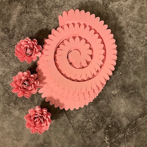 Pode incluir: Um conjunto de tr&ecirc;s flores de papel rosa e uma espiral de papel rosa. A espiral &eacute; feita de v&aacute;rias camadas de papel, cada uma com uma borda festonada.