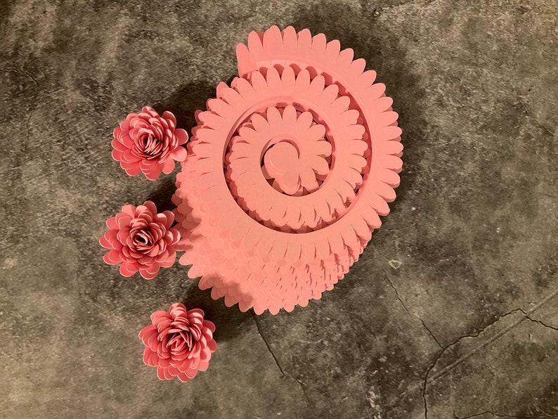 Pode incluir: Um conjunto de tr&ecirc;s flores de papel rosa e uma espiral de papel rosa. A espiral &eacute; feita de v&aacute;rias camadas de papel, cada uma com uma borda festonada.