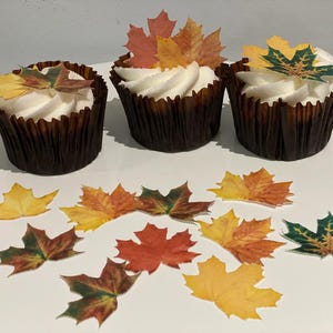 May include: Three cupcakes with white icing and autumn leaf decorations. The cupcakes are in brown paper cases. Scattered around the cupcakes are additional leaf decorations in shades of yellow, orange, and green.