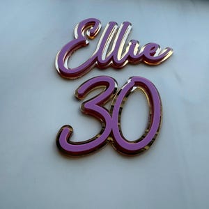 Può includere: Topper decorativo con il nome "Ellie" e il numero "30" in corsivo. Le lettere e i numeri sono di un rosa vibrante con un contorno dorato, perfetto per una festa di compleanno.