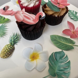 May include: Three cupcakes decorated with tropical themed edible images. The cupcakes are topped with pink flamingos, green palm leaves, white plumeria flowers, and pink hibiscus flowers.