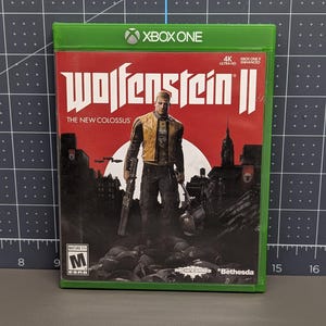 May include: A green plastic Xbox One video game case for Wolfenstein II: The New Colossus. The cover art features a character in a yellow jacket standing before a red sun and a dark cityscape. The ESRB M rating and Bethesda logo are visible.