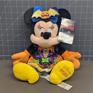 May include: A Minnie Mouse plush toy dressed for Halloween. She wears a purple and orange dress with a bow featuring a pumpkin and bat. Her yellow shoes are embroidered with "Disney 2023". A tag reads "Happy Halloween".