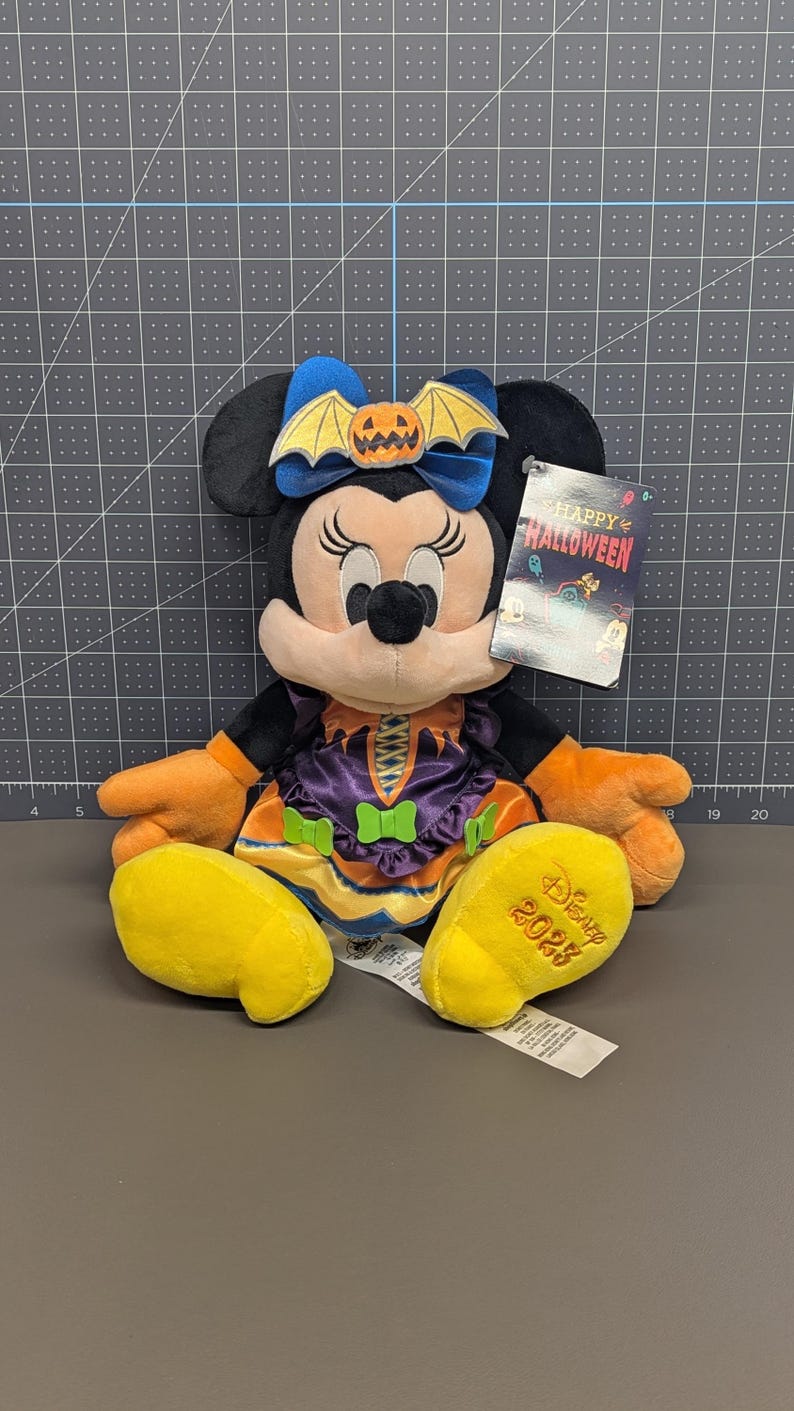 May include: A Minnie Mouse plush toy dressed for Halloween. She wears a purple and orange dress with a bow featuring a pumpkin and bat. Her yellow shoes are embroidered with "Disney 2023". A tag reads "Happy Halloween".