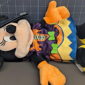 May include: A plush Minnie Mouse doll in a Halloween costume. The doll features a black head with large ears, a witch hat, and a bow with a pumpkin bat design. She wears a purple and orange dress with green bow accents and orange gloves and yellow shoes.