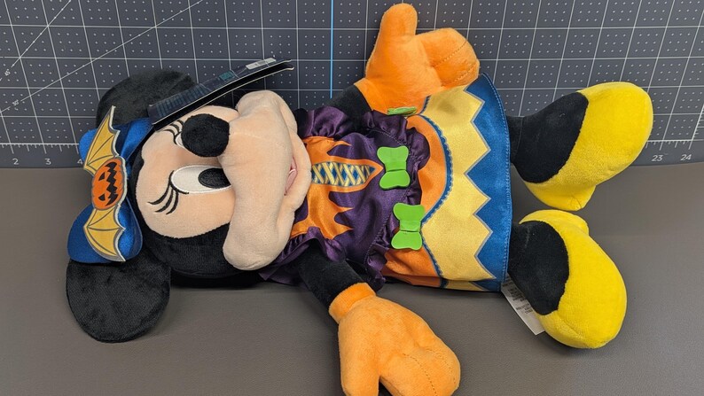 May include: A plush Minnie Mouse doll in a Halloween costume. The doll features a black head with large ears, a witch hat, and a bow with a pumpkin bat design. She wears a purple and orange dress with green bow accents and orange gloves and yellow shoes.