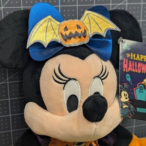 May include: A plush Minnie Mouse doll with a Halloween theme. She has black ears, a blue bow with a pumpkin bat, and a tag that says "Happy Halloween." The doll is wearing an orange and purple outfit.